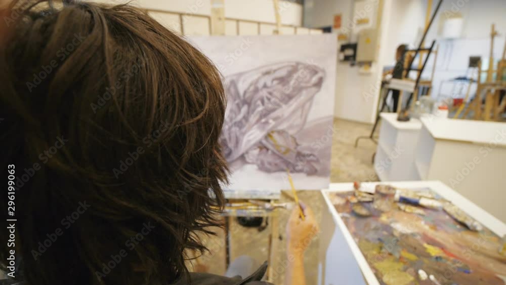 Male hispanic artist in black leather jacket paints oil still-life on canvas sitting on chair in art studio. Over the shoulder shot.