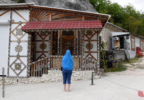 A woman with a covered head stands and admires the Orthodox decorations of the building. The building is decorated with shells and colorful pebbles.