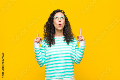 young pretty woman feeling awed and open mouthed pointing upwards with a shocked and surprised look against orange wall photo