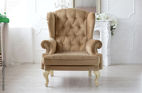 Soft beige armchair stands in the center of the white room with a fireplace. Vintage furniture. Cozy interior © Oksana