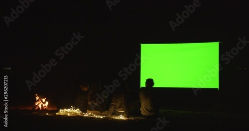 The company of young friends watching a movie on the beach by the fire. Movie night. Cinema under the open sky. Green screen.