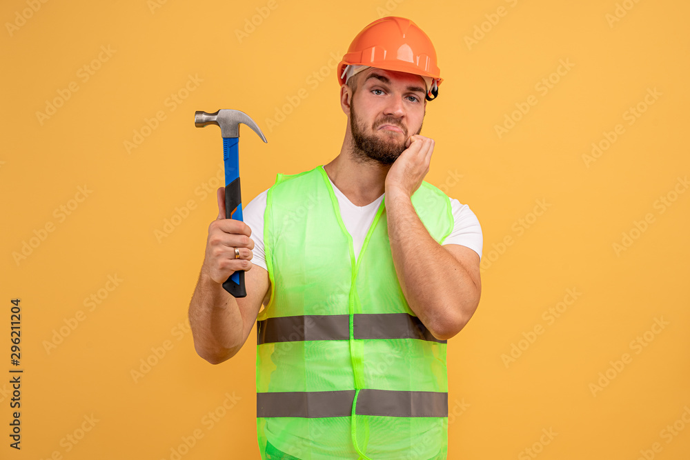 Home renovation concept. Indignant builder holds hammer, does repairing, wears building helmet, has labour work, looks with puzzlement at camera, isolated on yellow background. Handyman with tool Home renovation concept. Indignant builder holds hammer, does repairing, wears building helmet, has labour work, looks with puzzlement at camera, isolated on yellow background. Handyman with tool