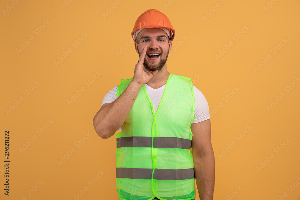 Mysterious surprised labourer worker looks with bugged eyes, whispers something, keeps palm near opened mouth, wears hardhat and uniform, involved in repairing or building. Handyman at factory