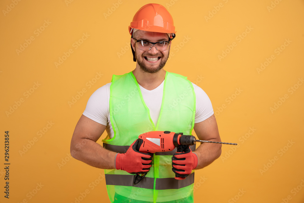 Bearded builder in helmet, reflective vest, glasses and gloves holding a drill in his hands. Isolated on yellow background. Yes, I did