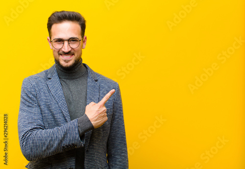 young handsome man smiling cheerfully, feeling happy and pointing to the side and upwards, showing object in copy space against orange wall