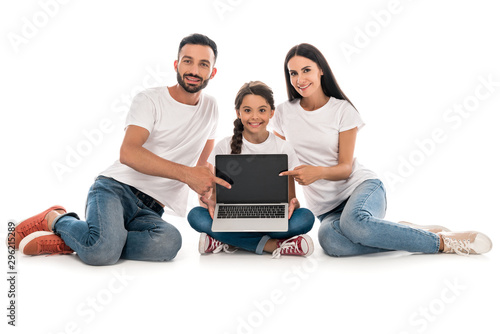 cheerful parents in white t-shirts pointing with fingers at laptop with blank screen near daughter isolated on white