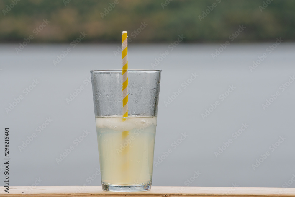 A tall glass of homemade lemonade with a yellow and white stripped paper straw in the glass. The glass is sitting on a table with the ocean behind it. A tall glass of homemade lemonade with a yellow and white stripped paper straw in the glass. The glass is sitting on a table with the ocean behind it.