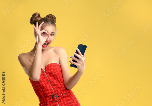 Vászonkép Happy girl holding a smartphone and showing okay sign wearing red dress on yello