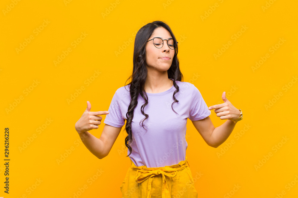 young pretty latin woman looking proud, positive and casual pointing to chest with both hands against orange wall - obrazy, fototapety, plakaty 