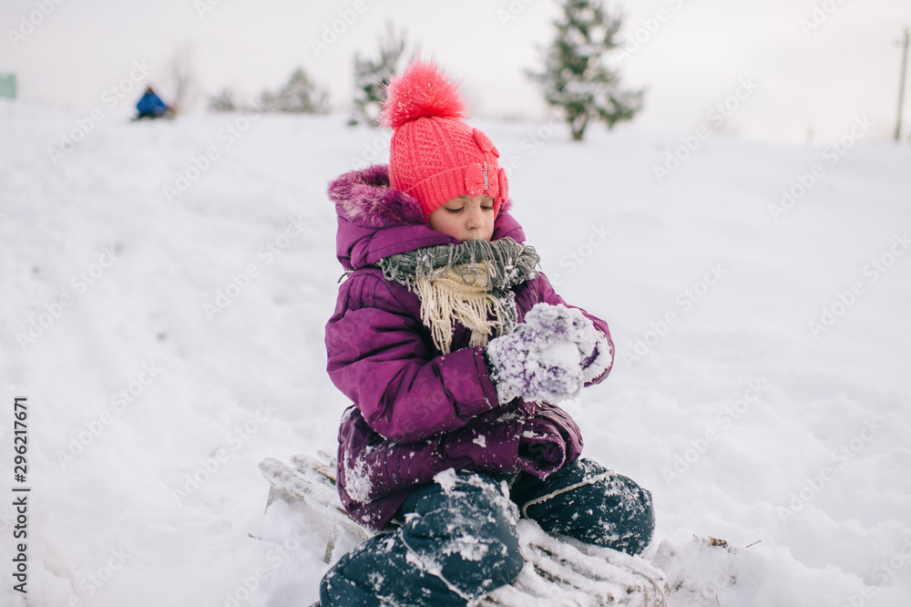 Little girl covered in snow sitting on sled with snobal in her hands in winter day. Happy child  playing with snow outdoors.