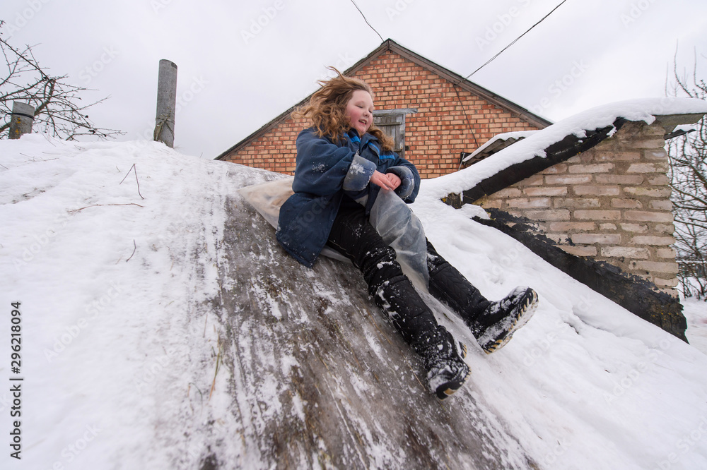Cheerful girl using plastic bag as a sled and riding down from cellar with icey trek in front of brick village house in winter day. Cheerful girl using plastic bag as a sled and riding down from cellar with icey trek in front of brick village house in winter day.