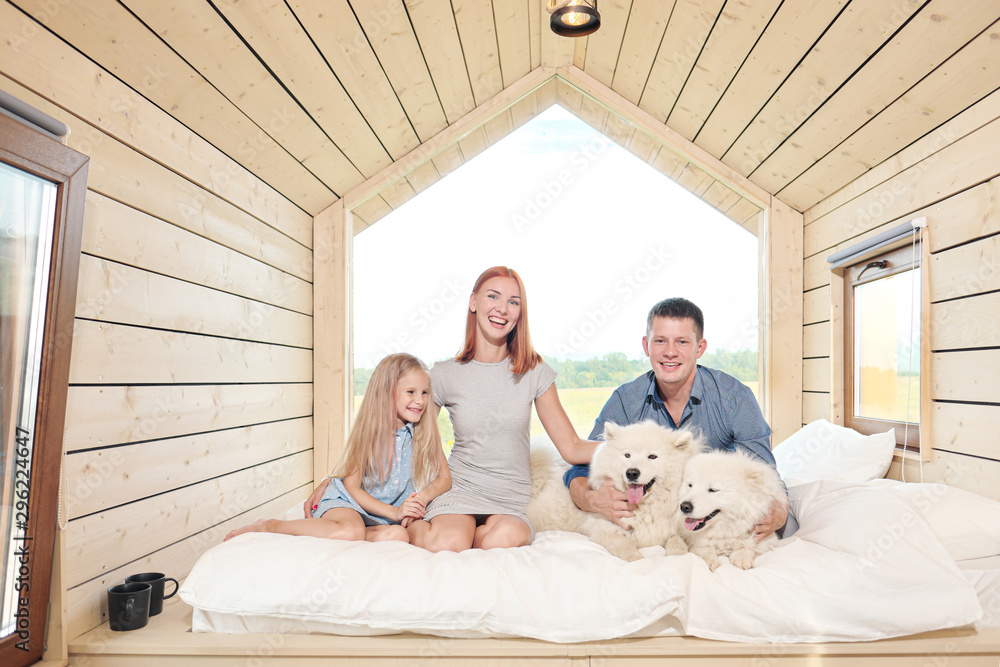 Young Caucasian family couple with baby daughter in a small modern rustic house with a large window. Lying on the bed, hugging, playing and looking out the window. Two Samoyeds. Weekend vacation.