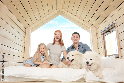 Fototapeta Naklejka Na Ścianę i Meble -  Young Caucasian family couple with baby daughter in a small modern rustic house with a large window. Lying on the bed, hugging, playing and looking out the window. Two Samoyeds. Weekend vacation.