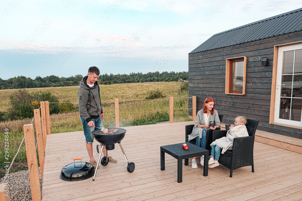 A young Caucasian family couple with a baby daughter sits on the terrace in chairs and drinks hot drinks. Against the background of a small modern rustic wooden house. Father is cooking barbecue.