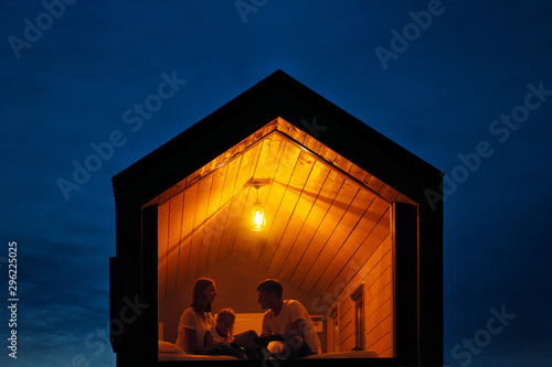 A family of three is sitting on a bed by the window, reading a book and talking. View of the outside of the house. Night blue sky. Orange warm light from an incandescent lamp.
