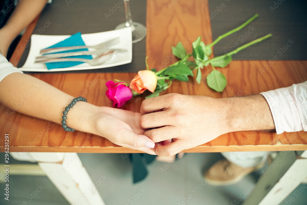 Young couple sitting at wooden table of indoor restaurant and holding hands while celebrating Valentines Day, Anniversary or wedding , two roses defocused - Image