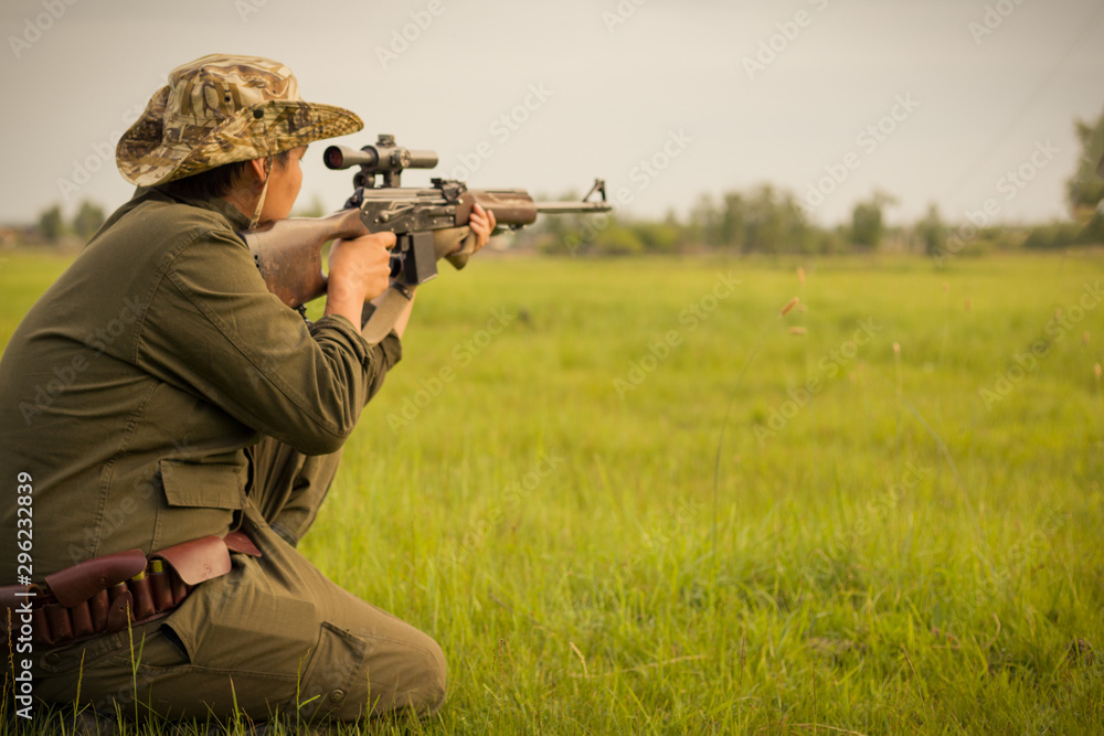 A male hunter with a gun while sitting takes aim at a forest. The concept of a successful hunt, an experienced hunter. Hunting the summer season. The hunter has a rifle and a hunting uniform. Image. A male hunter with a gun while sitting takes aim at a forest. The concept of a successful hunt, an experienced hunter. Hunting the summer season. The hunter has a rifle and a hunting uniform. Image.