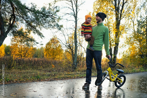Father and his tiddler son walking in the park on good autumn day. Dad holding kid in one hand and child's bicycle in another. photo