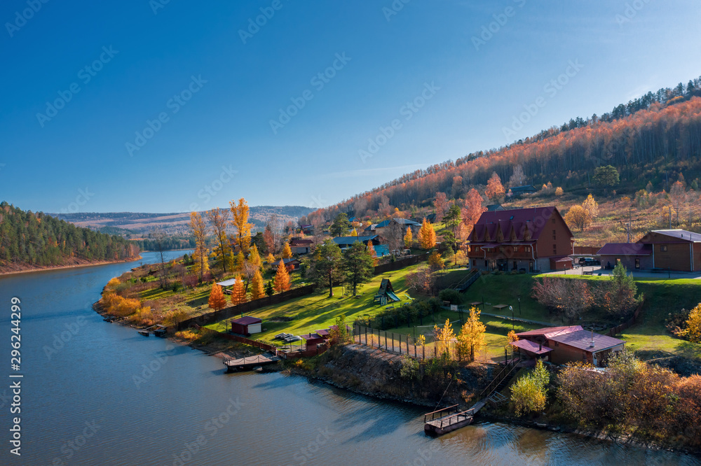 Aerial view; drone flying around little village on riverside; old buildings in autumn landscape, colorful forest; orange yellow foliage of birch trees; beautiful countryside near pond, Porogi, Ural