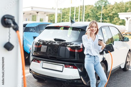 Young adult woman standing on parking, charging electric car, talking on smartphone
