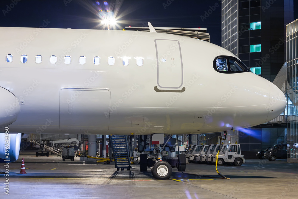 The front part of the white passenger airplane on the airport apron near the air bridge and terminal at night The front part of the white passenger airplane on the airport apron near the air bridge and terminal at night