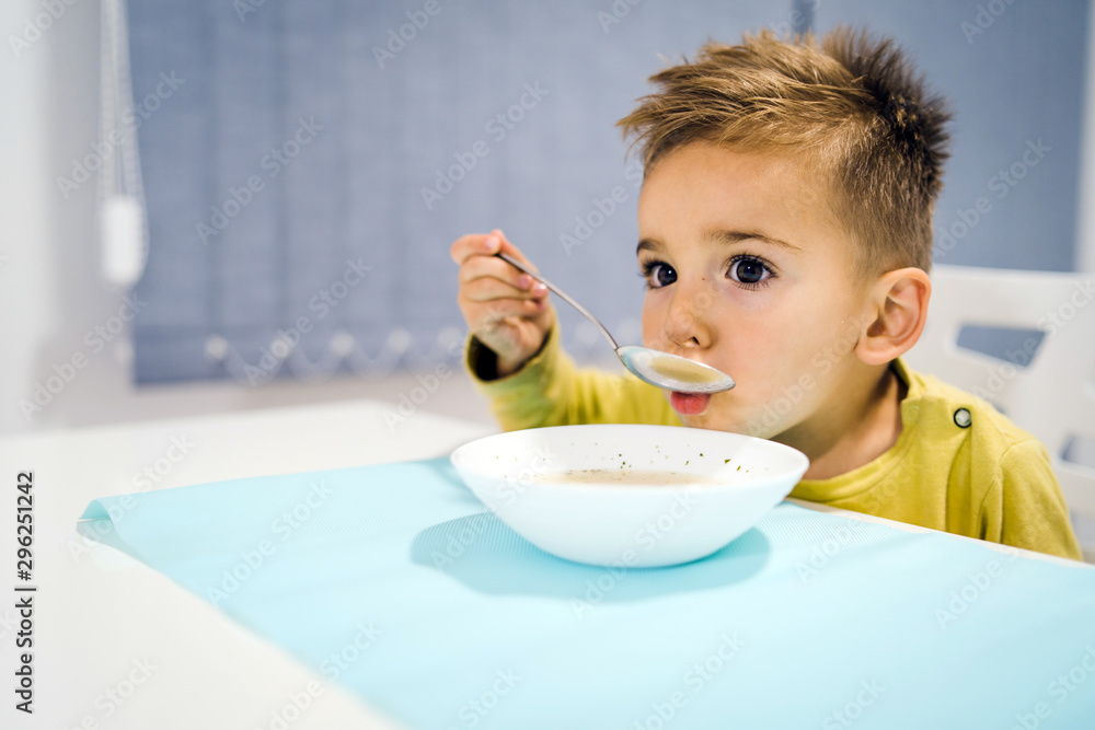 Portrait of small boy child eating soup meal or breakfast having lunch by the table at home with spoon white kindergarten Portrait of small boy child eating soup meal or breakfast having lunch by the table at home with spoon white kindergarten