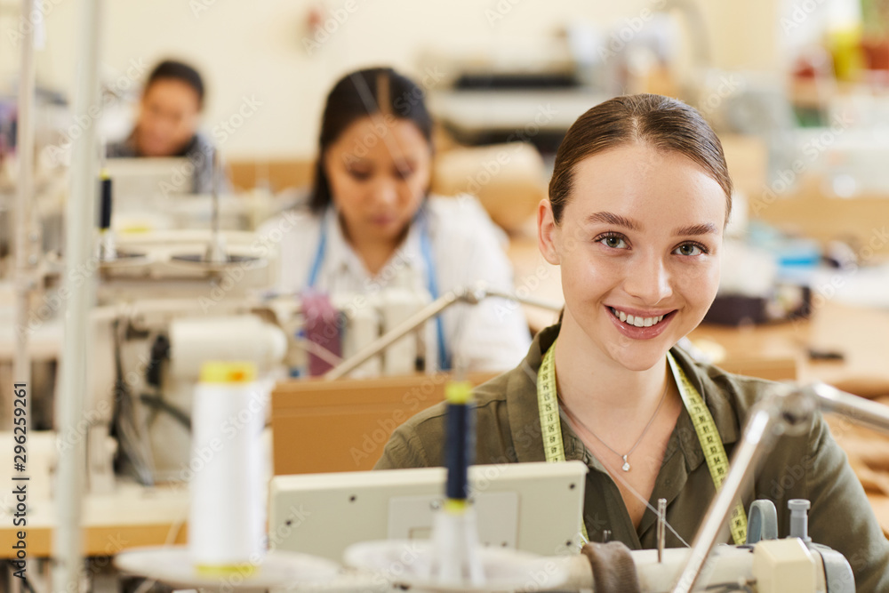 Portrait of young tailor smiling at camera while sewing on sewing machine with her colleagues in the background in workshop Portrait of young tailor smiling at camera while sewing on sewing machine with her colleagues in the background in workshop