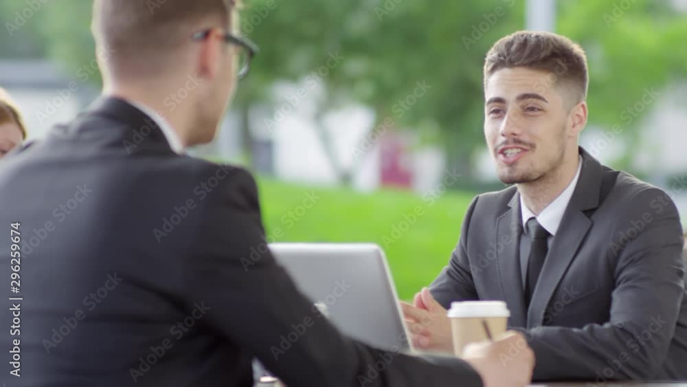 Tracking medium shot of young businessman smiling while talking to unrecognizable colleague while having lunch break in outdoor cafe Tracking medium shot of young businessman smiling while talking to unrecognizable colleague while having lunch break in outdoor cafe