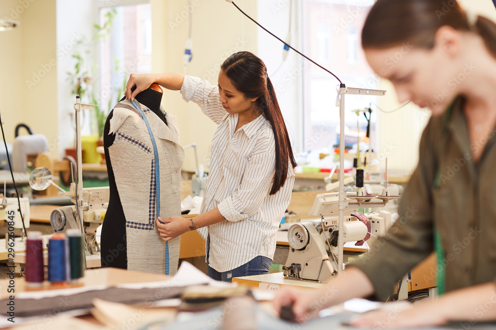 Obraz premium Asian young designer measuring suit with tape measure hanging on mannequin in workshop