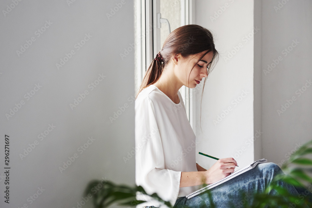 On the windowsill. Young brunette in the room with white walls and daylight that comes from the window