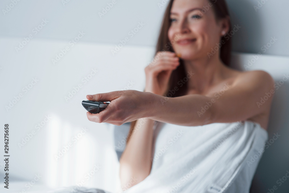 Focus on the remote controller. Pretty young woman sitting on the bed at morning time in her room Focus on the remote controller. Pretty young woman sitting on the bed at morning time in her room