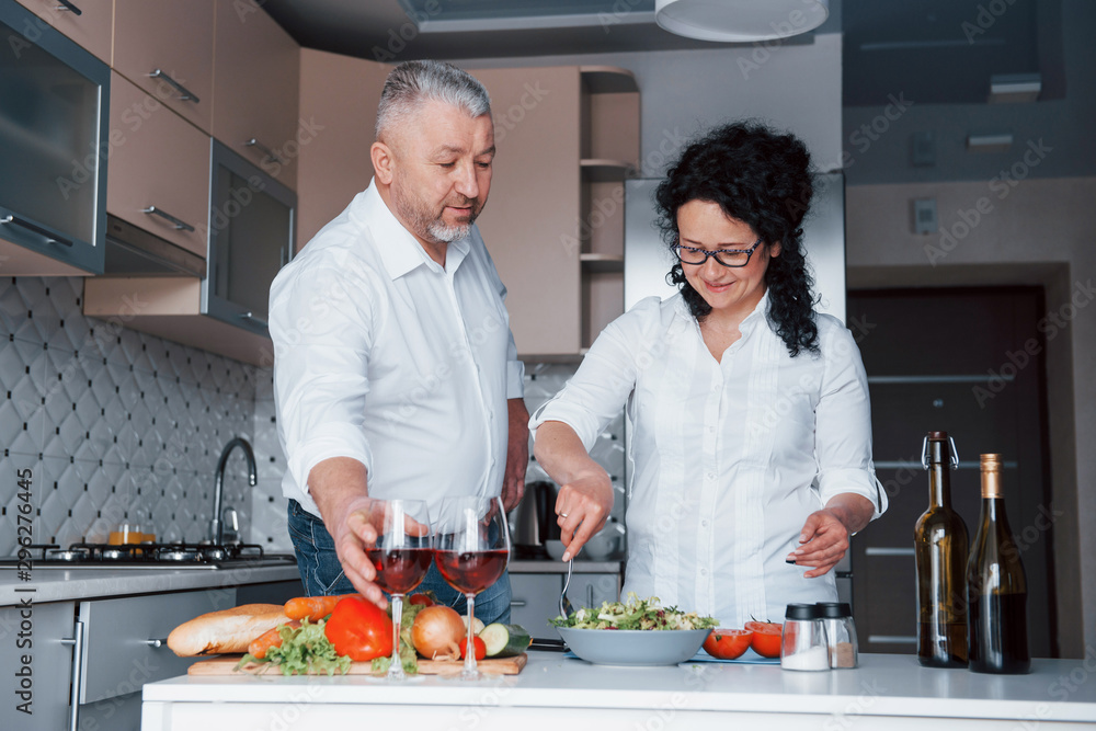Happy senior couple. Man and his wife in white shirt preparing food on the kitchen using vegetables Happy senior couple. Man and his wife in white shirt preparing food on the kitchen using vegetables