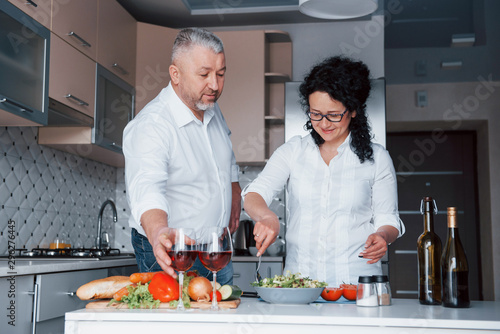 Happy senior couple. Man and his wife in white shirt preparing food on the kitchen using vegetables