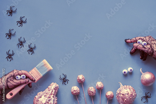 Creative Halloween flat lay on blue-grey paper background with copy-space. Frame made of hand in black mesh gloves imitating spider with candy eyes, holding paint brush and decorative pumpkins.