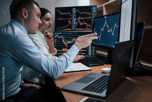 Man talks and shows on the numbers on monitor with hand. Team of stockbrokers are having a conversation in a office with multiple display screens