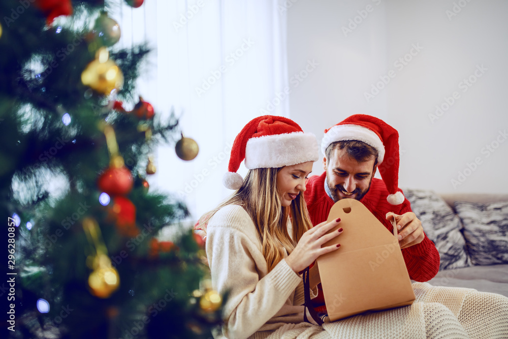 Obraz premium Adorable smiling caucasian couple with santa hats on heads siting on sofa in living room and opening gift. In foreground is christmas tree.