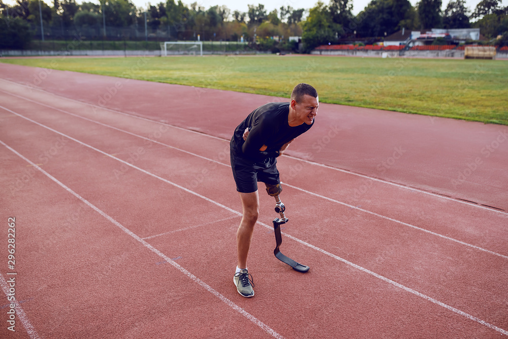 Sporty caucasian man with artificial leg standing on running track and having pain in stomach.