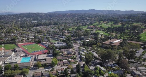 Aerial of houses in suburbs with college football ground and lush green golfing ground on a sunny day with clear blue sky and mountains in the background