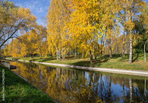 Autumn park with yellow leaves and trees with a river and reflection in the water and blue sky.