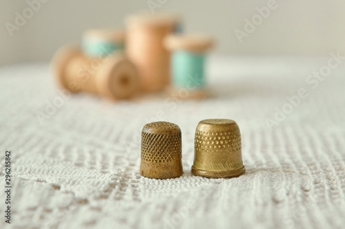 Two vintage thimbles close-up, in the background old wooden bobbins with sewing threads. Are on the tablecloth antique hand knit lace.