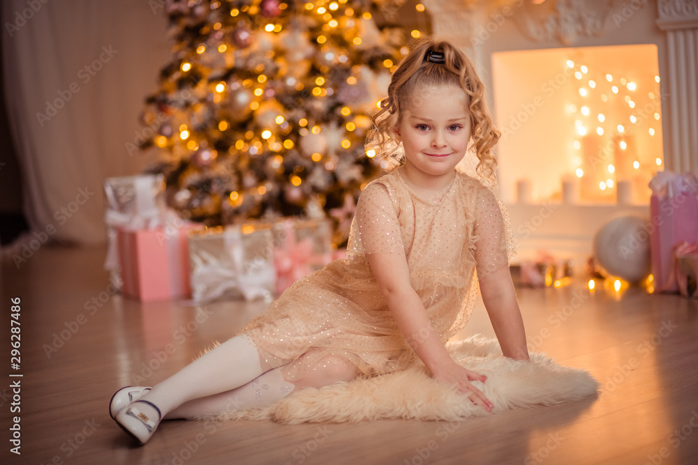 little cute girl in a beautiful interior sits on the background of the fireplace and New Year tree in anticipation of the holiday. lanterns, lights and gifts are visible in the background