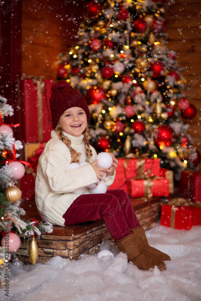 cheerful cute girl In a light knitted sweater and red hat sits on the steps of a wooden house in anticipation of the New Year holiday, in her hands she holds snowballs and has fun cheerful cute girl In a light knitted sweater and red hat sits on the steps of a wooden house in anticipation of the New Year holiday, in her hands she holds snowballs and has fun