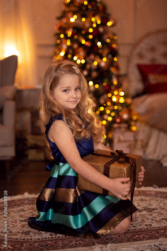 A cute girl with blond hair with a gift in her hands is sitting waiting for the holiday against the background of the Christmas tree in the lights. A cute girl with blond hair with a gift in her hands is sitting waiting for the holiday against the background of the Christmas tree in the lights.