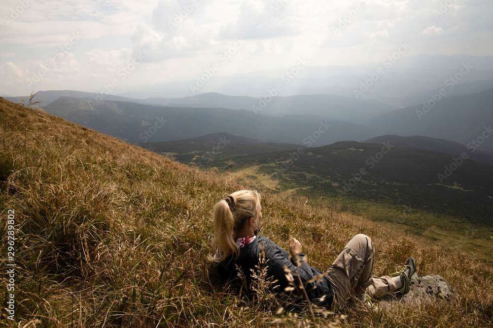 Fototapeta premium Tourist girl resting in the mountains. Tourist girl lying on the grass in the mountains. Stylish traveler woman lying on grass and relaxing in mountains. Travel concept.