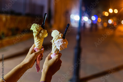 In women's hands two tubes of ice cream with different flavors, strawberries, banana, cream, on background of night street of city with blurred color lights.Young girl holding ice cream cones