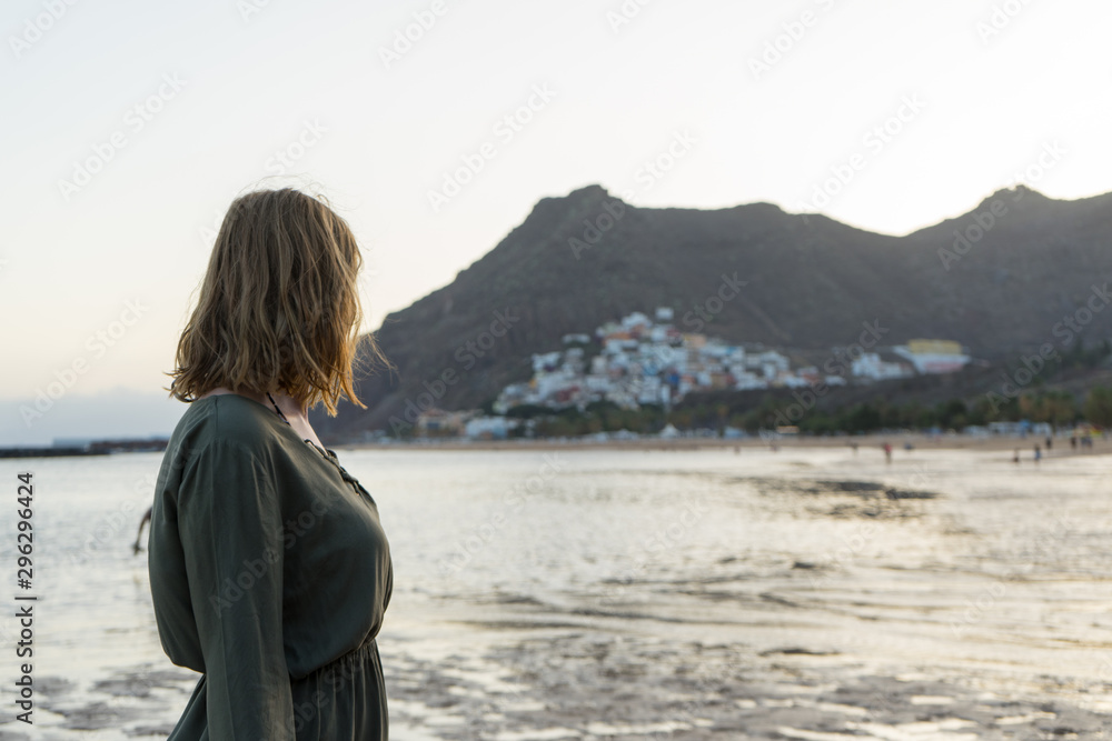 Spiritual adolescent in green dress admire the sunset and the sea town on a hill in Tenerife. Blonde romantic woman look the paradise in her vacation in calm