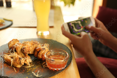 Woman playing game using smartphone at table in cafe, focus on tasty BBQ wings