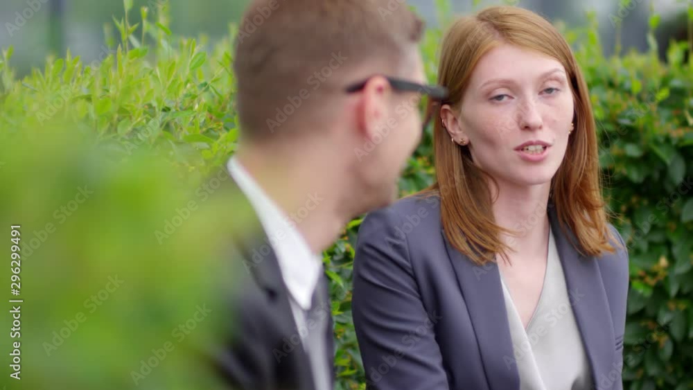 Medium shot of young Caucasian businesswoman talking to male colleague while relaxing in park during break from work Medium shot of young Caucasian businesswoman talking to male colleague while relaxing in park during break from work
