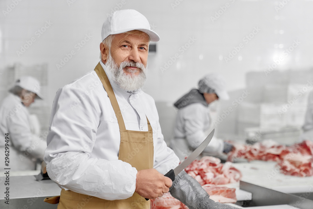 Elderly man working on butchery, cutting meat with knife.