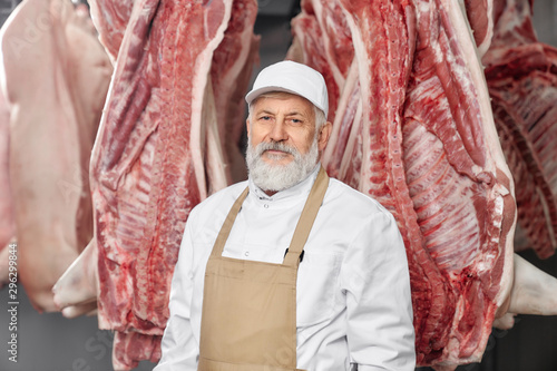 Professional butcher in uniform standing near pork carcasses photo